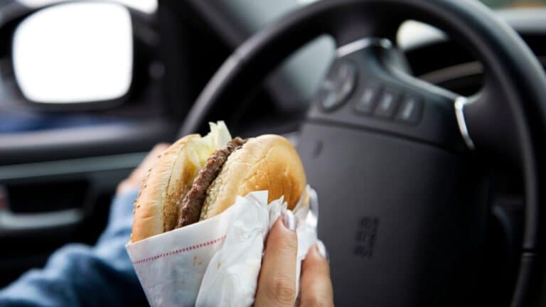 On-the-Go Deliciousness: Close-Up of Man Enjoying a Hamburger While Driving. Cheeseburger, fast food sandwich, driving while eating, eating in the car.