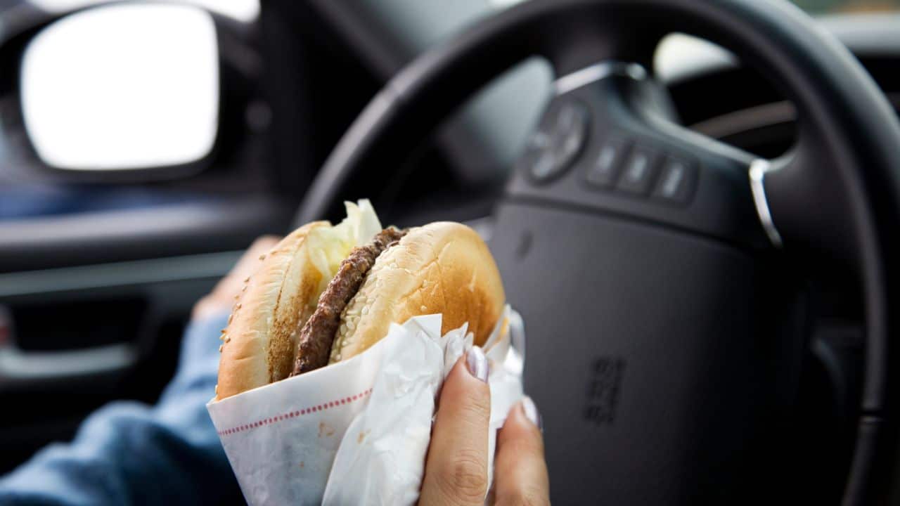 On-the-Go Deliciousness: Close-Up of Man Enjoying a Hamburger While Driving. Cheeseburger, fast food sandwich, driving while eating, eating in the car.