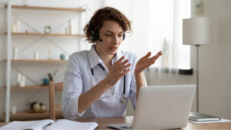 Female general practitioner wears white coat and headset speaking videoconferencing on laptop computer using online video call consultation app. Remote medical help for distance patient, telemedicine.