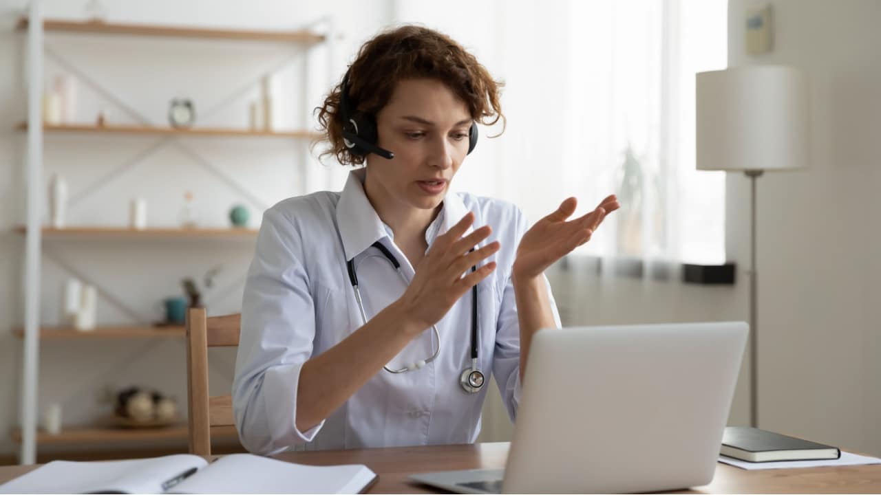Female general practitioner wears white coat and headset speaking videoconferencing on laptop computer using online video call consultation app. Remote medical help for distance patient, telemedicine.