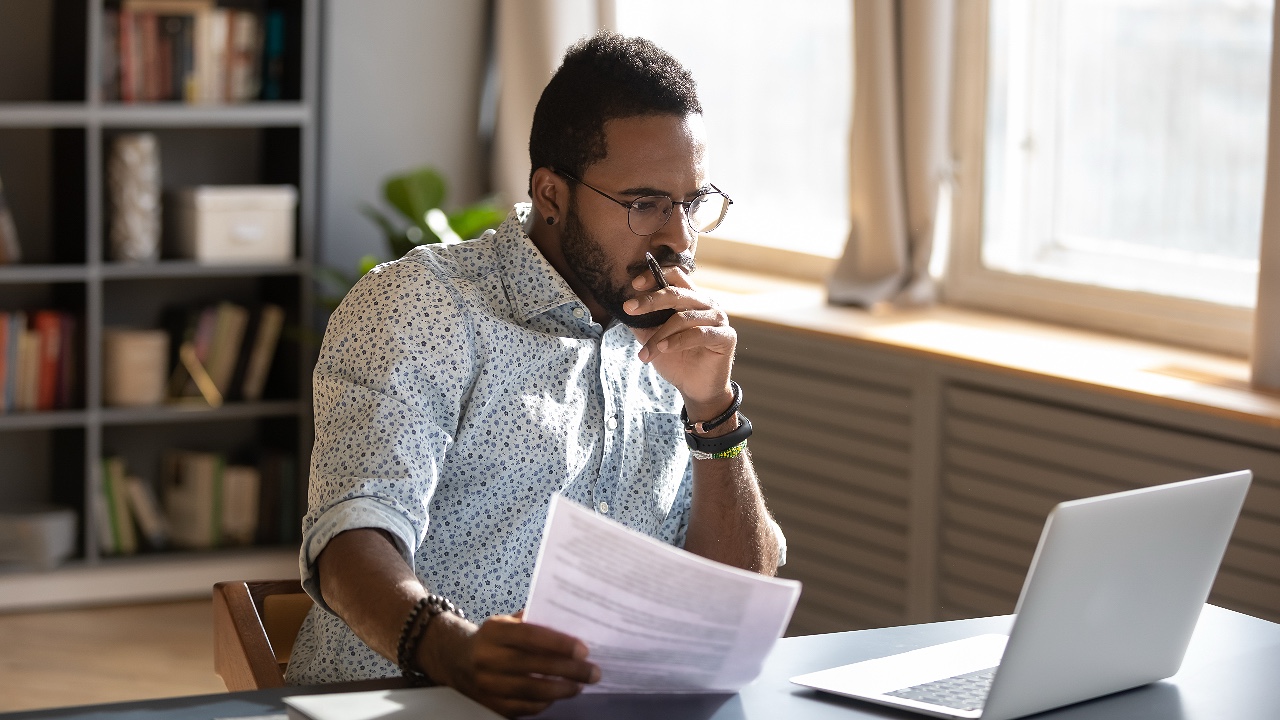 Focused businessman accountant analyst holding documents looking at laptop computer screen doing online trade market tech research thinking working sit at home office desk