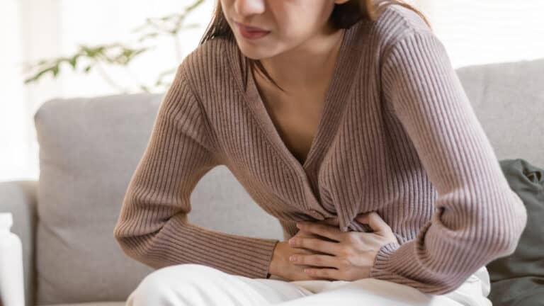 A young woman in a tan sweater and white trousers clutching her stomach in pain.