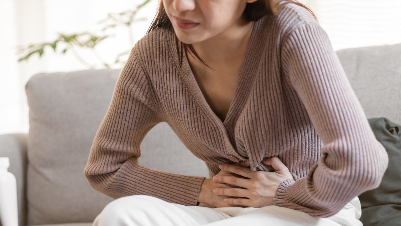 A young woman in a tan sweater and white trousers clutching her stomach in pain.