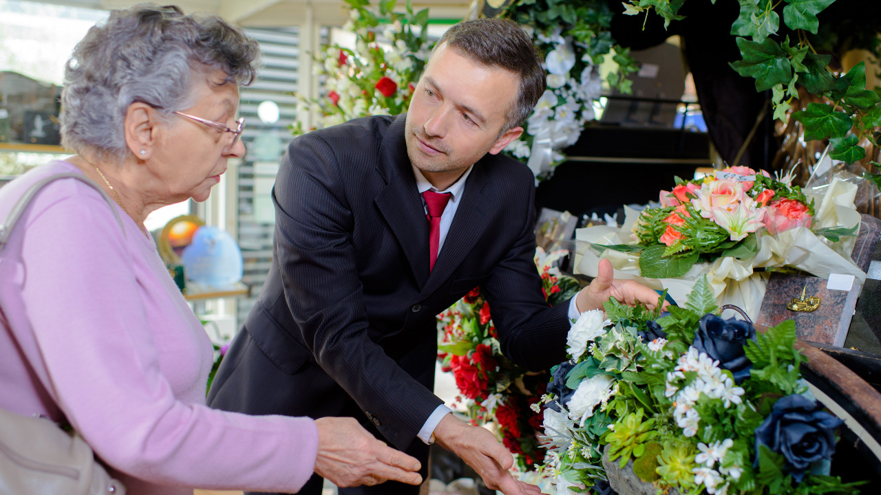 A funeral director assists a woman next to a casket and flower arrangement