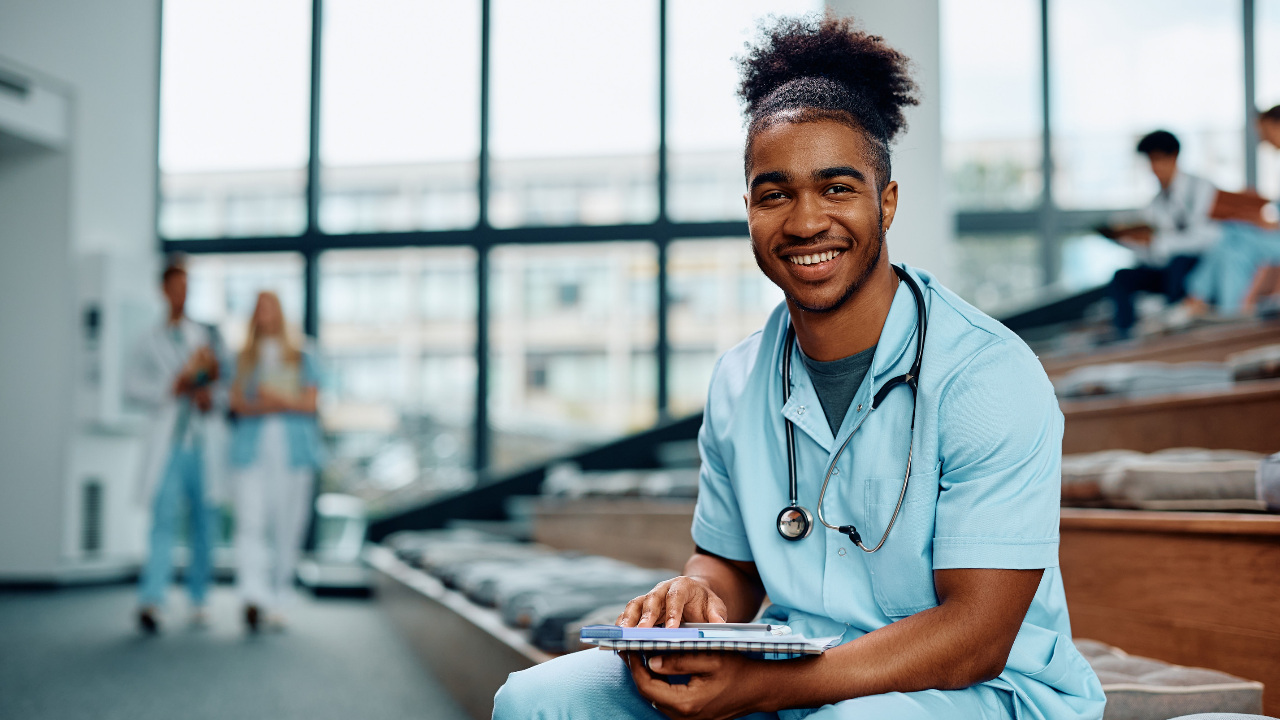 A young man smiling in a college lecture room while wearing scrubs and stethoscope
