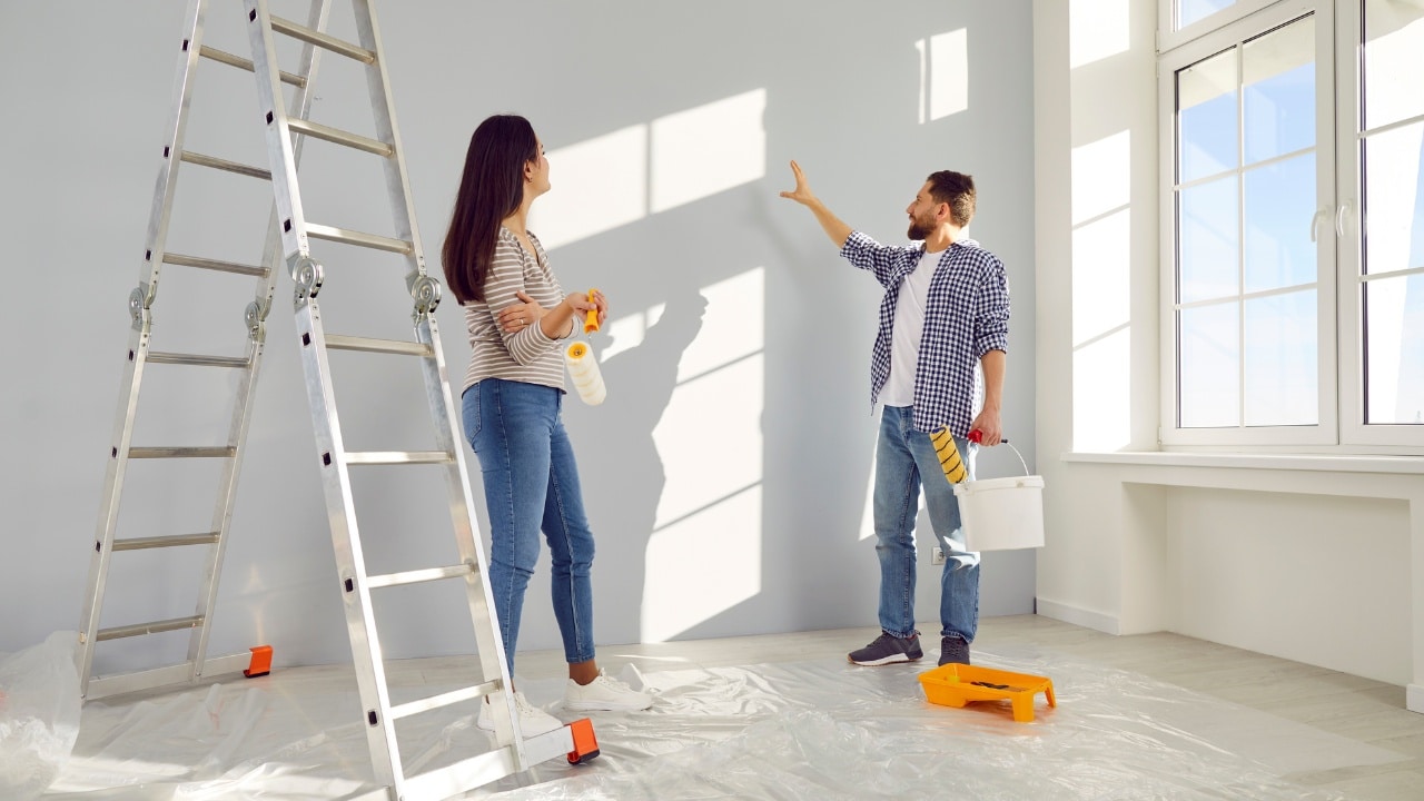 A couple painting and planning out their new living room interior design together. They both hold paint brushes while other items surround them.