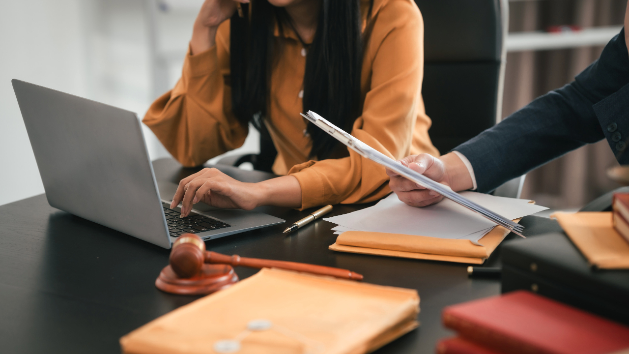 Two people sitting at a table surrounded by documents, laptops, with a gavel