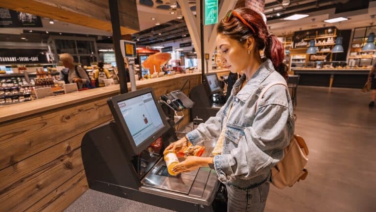 A young woman scans and pays for a bottle of juice at an automated self-checkout service terminal.