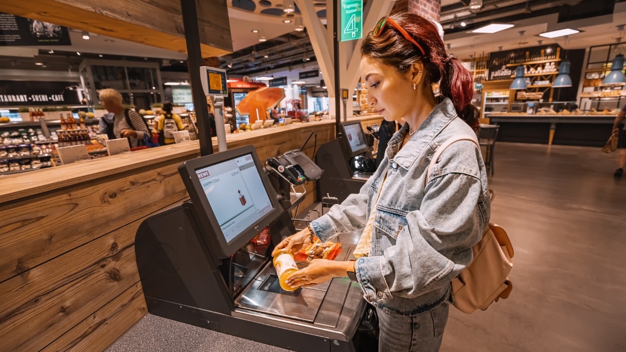 A young woman scans and pays for a bottle of juice at an automated self-checkout service terminal.