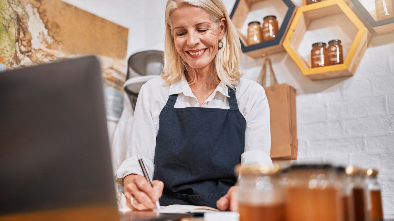 Laptop, small business and senior woman writing notes while doing inventory or online marketing. Happy, smile and elderly entrepreneur in a honey store working on admin or shop sales with a computer.