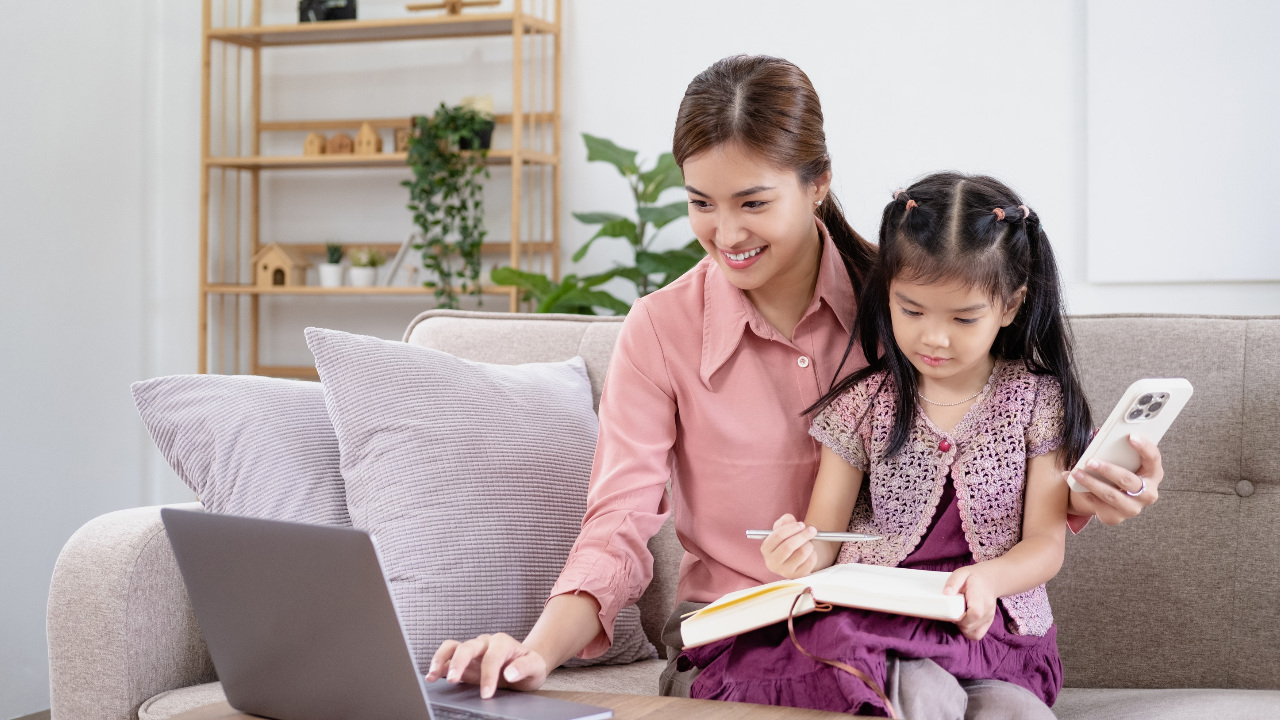 A mother with a phone in one hand and other hand on laptop keyboard holds her child while planning for the future