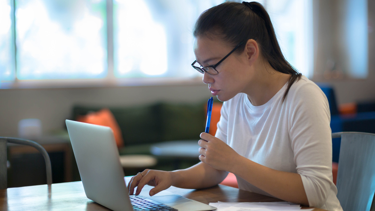 A woman sits with a laptop, taking notes on pen and paper