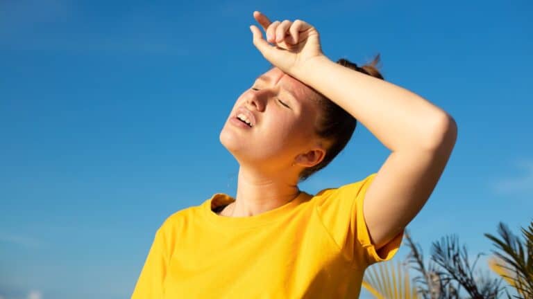 A young woman in a yellow tee shirt holds her arm against her forehead in an attempt to block out the sun and wipe sweat from her forehead.