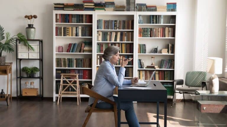 Side full length view focused mature senior businesswoman dictating audio message on cellphone, working on laptop in modern home office, recording voice message or holding loudspeaker conversation.
