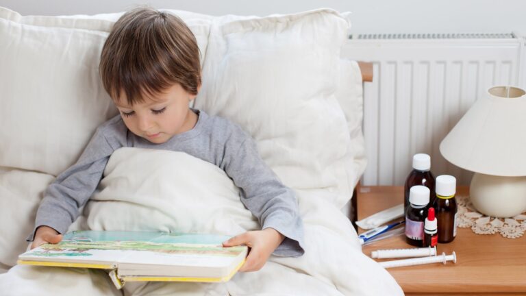 Sick child boy lying in bed with a fever, resting reading a book.