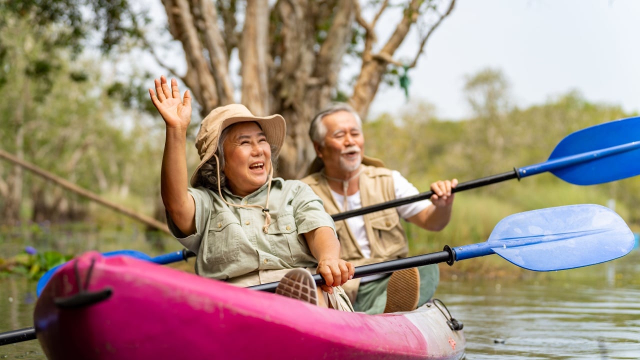 Asian senior couple kayaking together in the lake at mangrove forest on summer vacation. Retired elderly people man and woman have fun outdoor lifestyle travel nature and rowing a boat in the river.