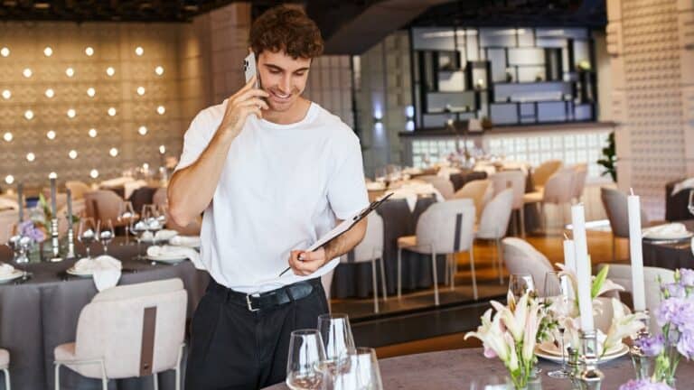 smiling event coordinator with clipboard talking on smartphone near festive tables in banquet hall, party planner.