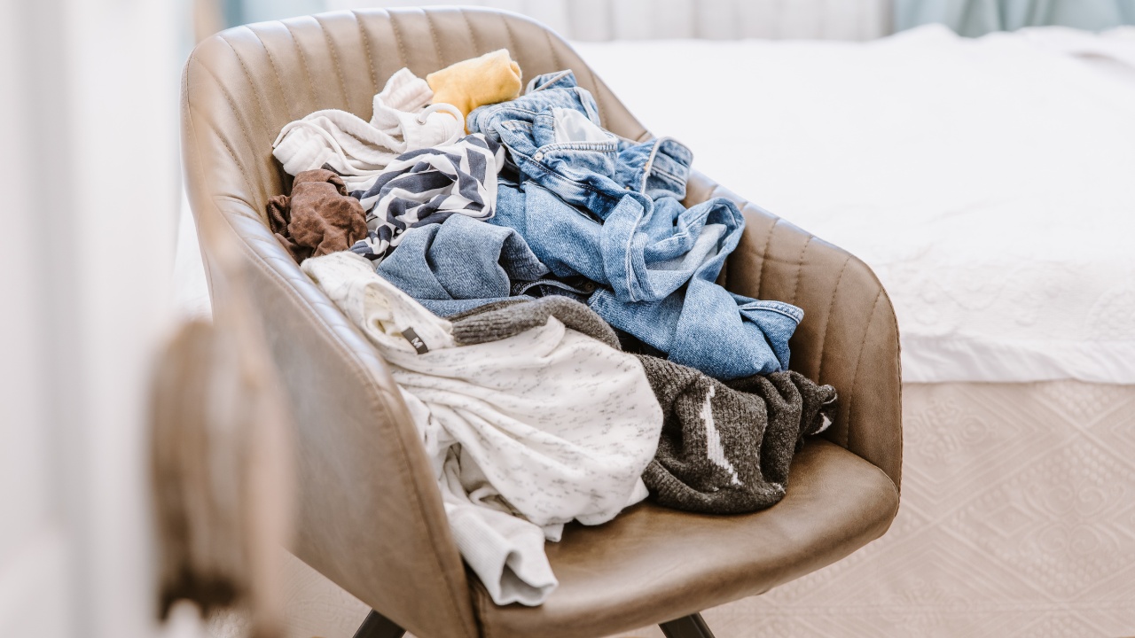 Pile of unfolded dirty clothes for laundry on a tan leather chair in a bedroom.