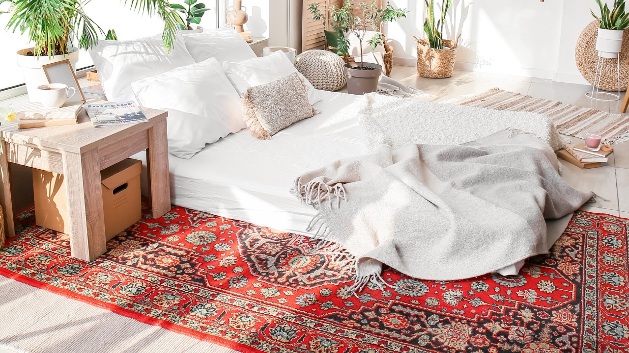 Interior of cozy bedroom with houseplants and vintage red rug underneath the bed. Wicker, bamboo and wooden furniture is scattered about the room. 