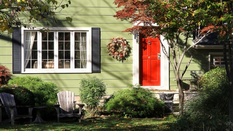 View of the exterior of a residential home, complete with light green siding, greenery, seating, shutters, decor and a bold red door.