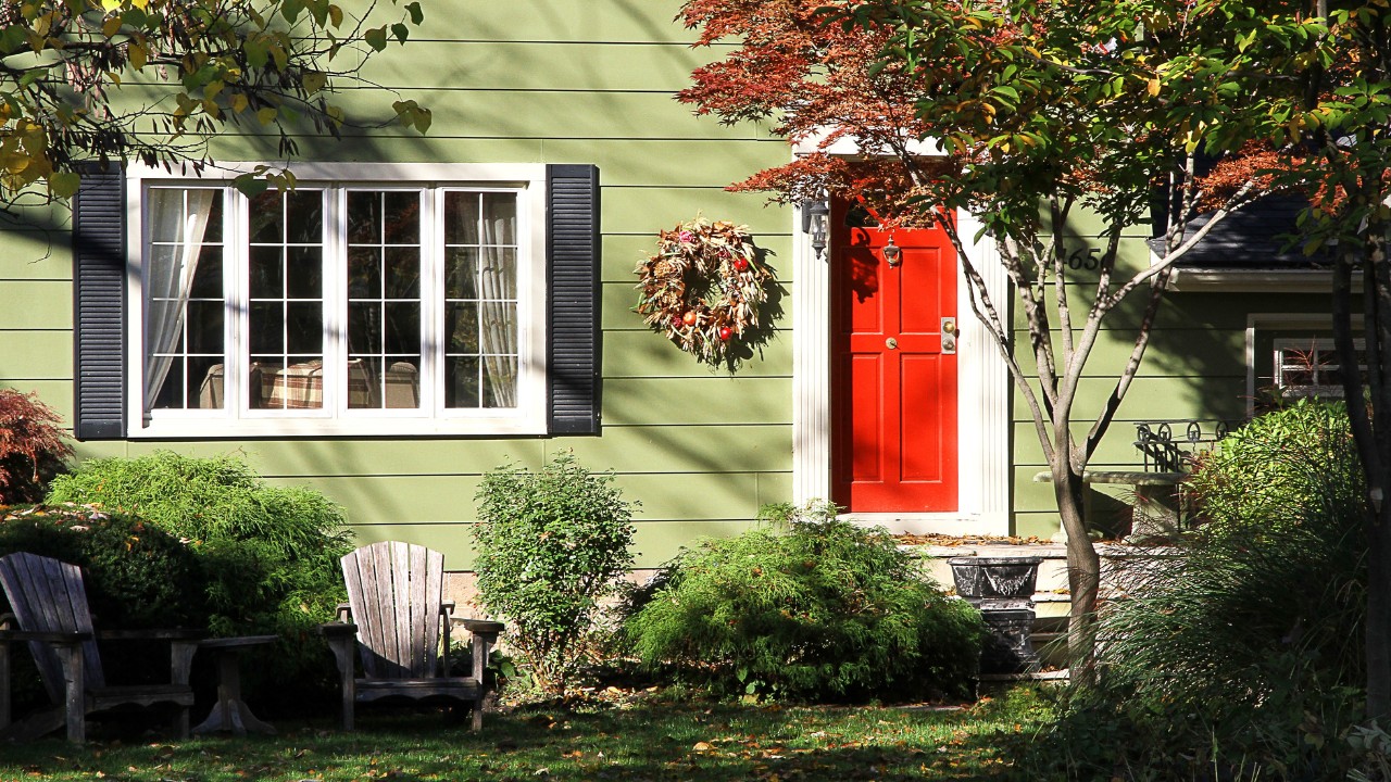 View of the exterior of a residential home, complete with light green siding, greenery, seating, shutters, decor and a bold red door.