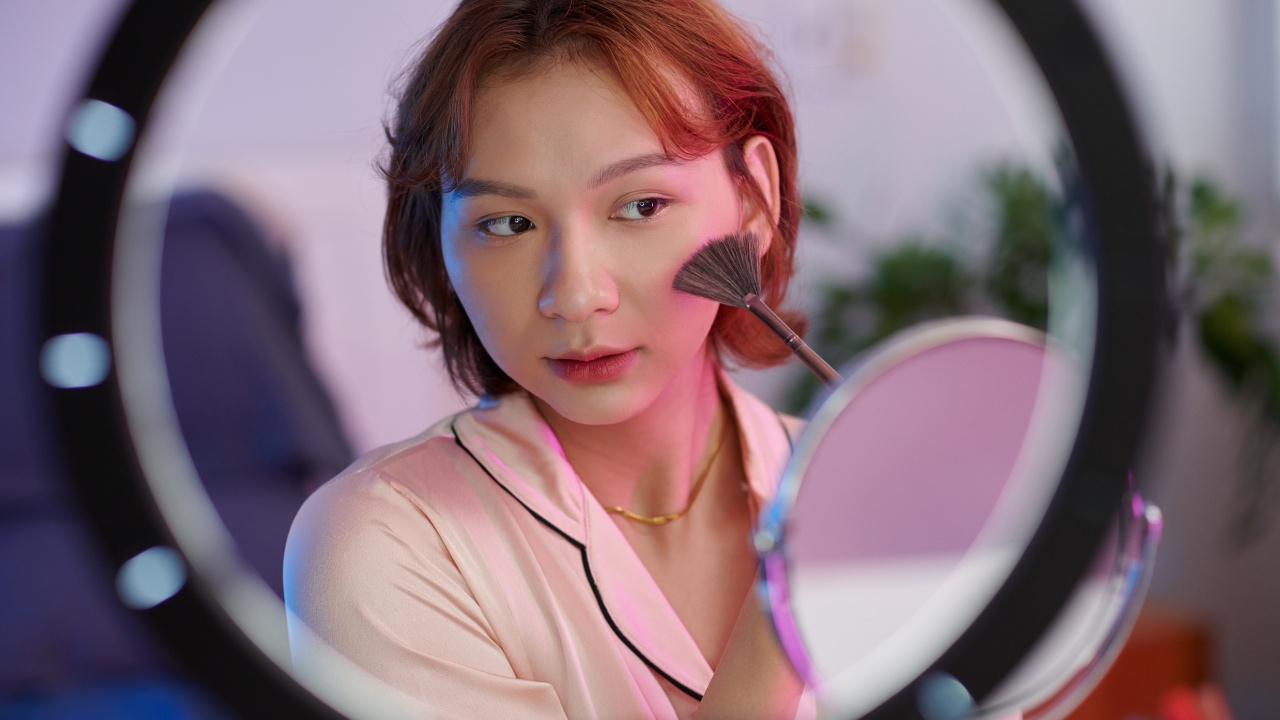 A young woman applying natural cheekbone highlight with a fan brush. There is a ring light and small mirror in front of her.
