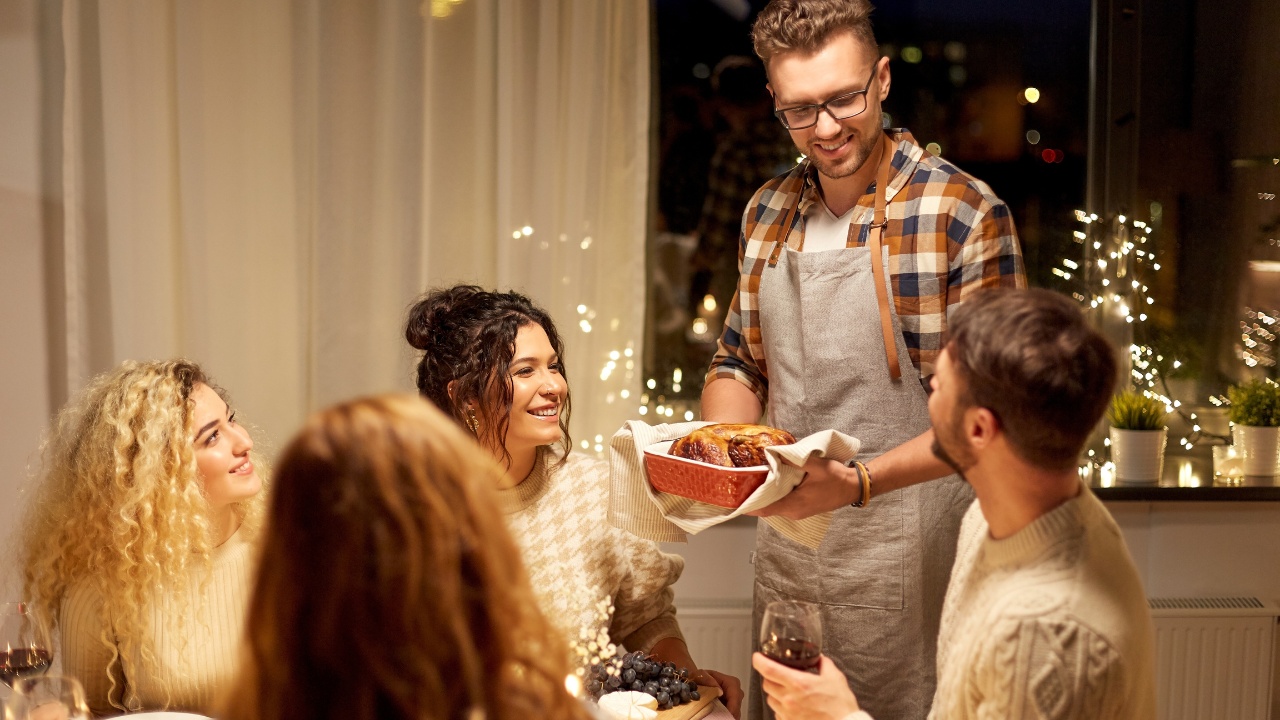A small group of people celebrating the holidays in a small living space. The room is decorated with lights, while the host serves the meal. The guests are seated around the table with wine and charcuterie.