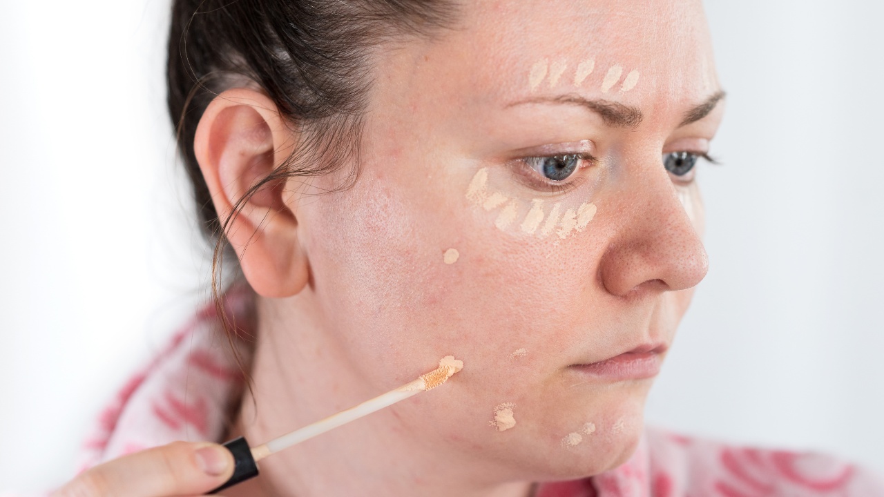 Young woman applies concealer makeup on her face with a small brush.