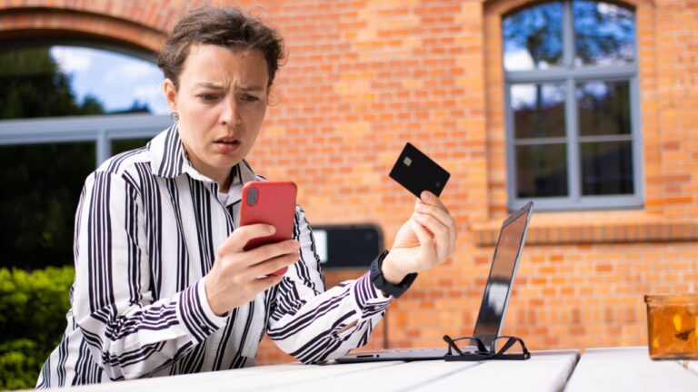 Confused business woman holding credit card looking at mobile phone sitting at table outdoors red brick building background Debt problems failed transaction money error contacting with scammer