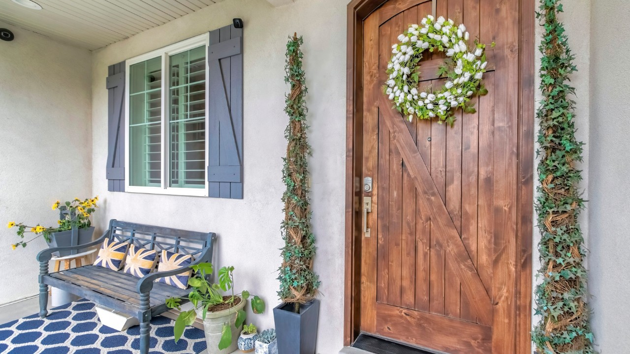 Panorama Porch of a house with wooden front door with digital key access. There are plants at the entrance and bench on a blue carpet near the window with fake shutter design.
