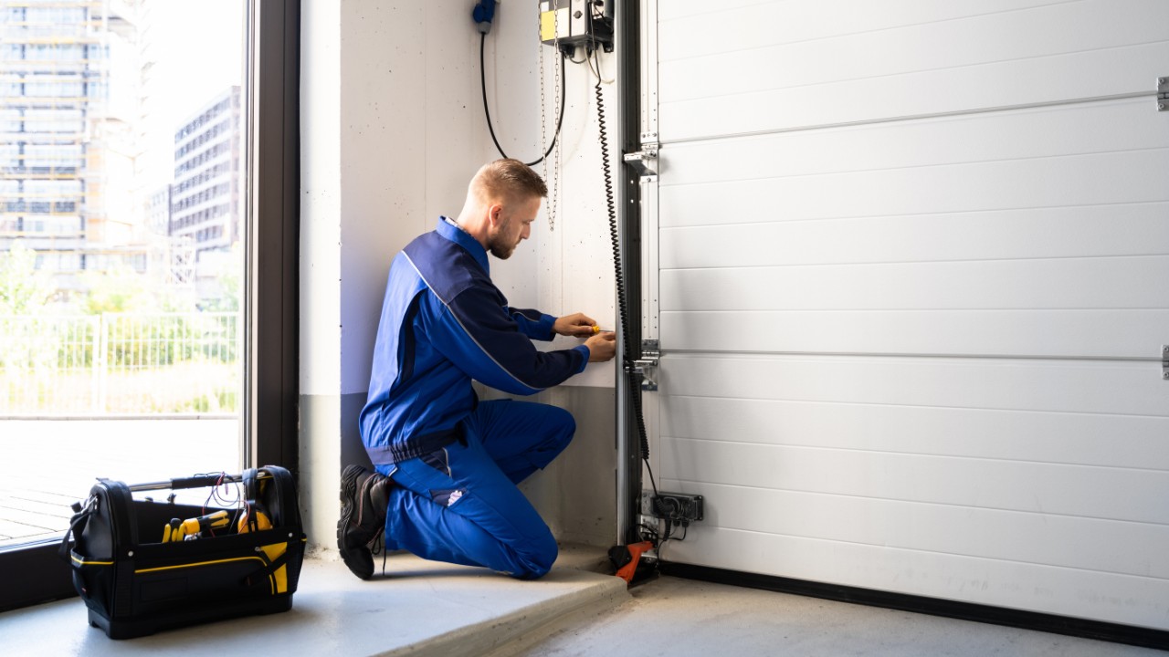 A male garage door technician installing a new unit at a residential home. Beside him rests a tool bag.
