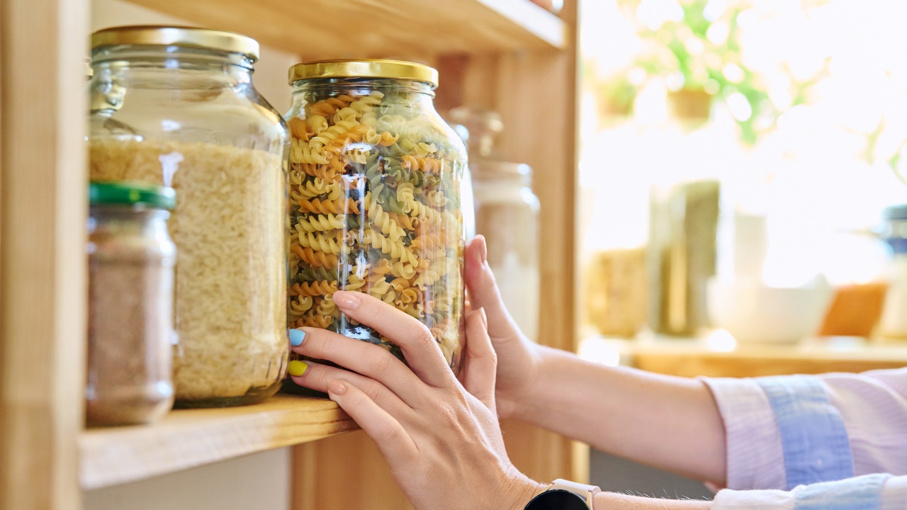 A feminine hand placing a large glass jar of dry stock pasta noodles in a pantry.