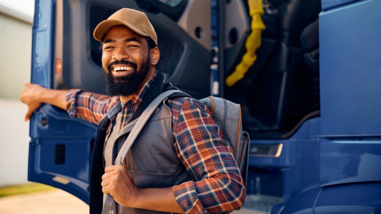 Male truck driver wearing flannel holding the driver's side door open to his large truck, smiling at the camera.