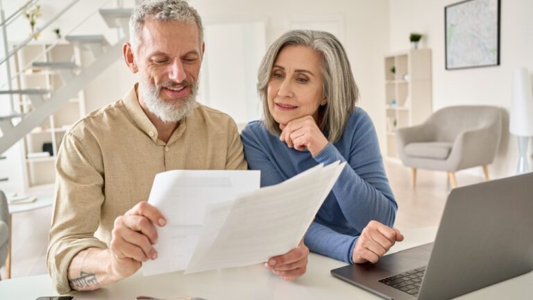 Middle aged senior old couple holding documents reading paper bills paying bank loan online, calculating pension fees, payments, taxes, planning family retirement money finances using laptop at home