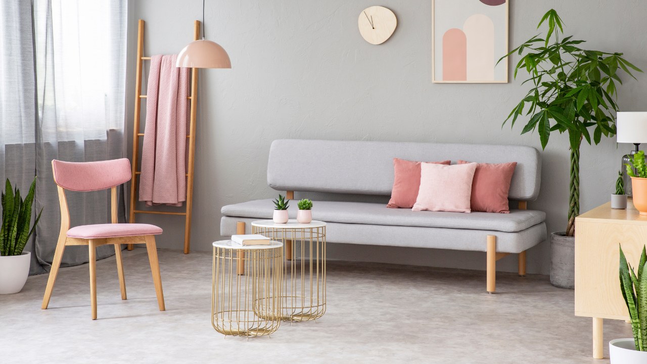 Real photo of lounge with dirty pink pillows, simple poster, pink chair, gold end tables with cacti and window with curtains. 