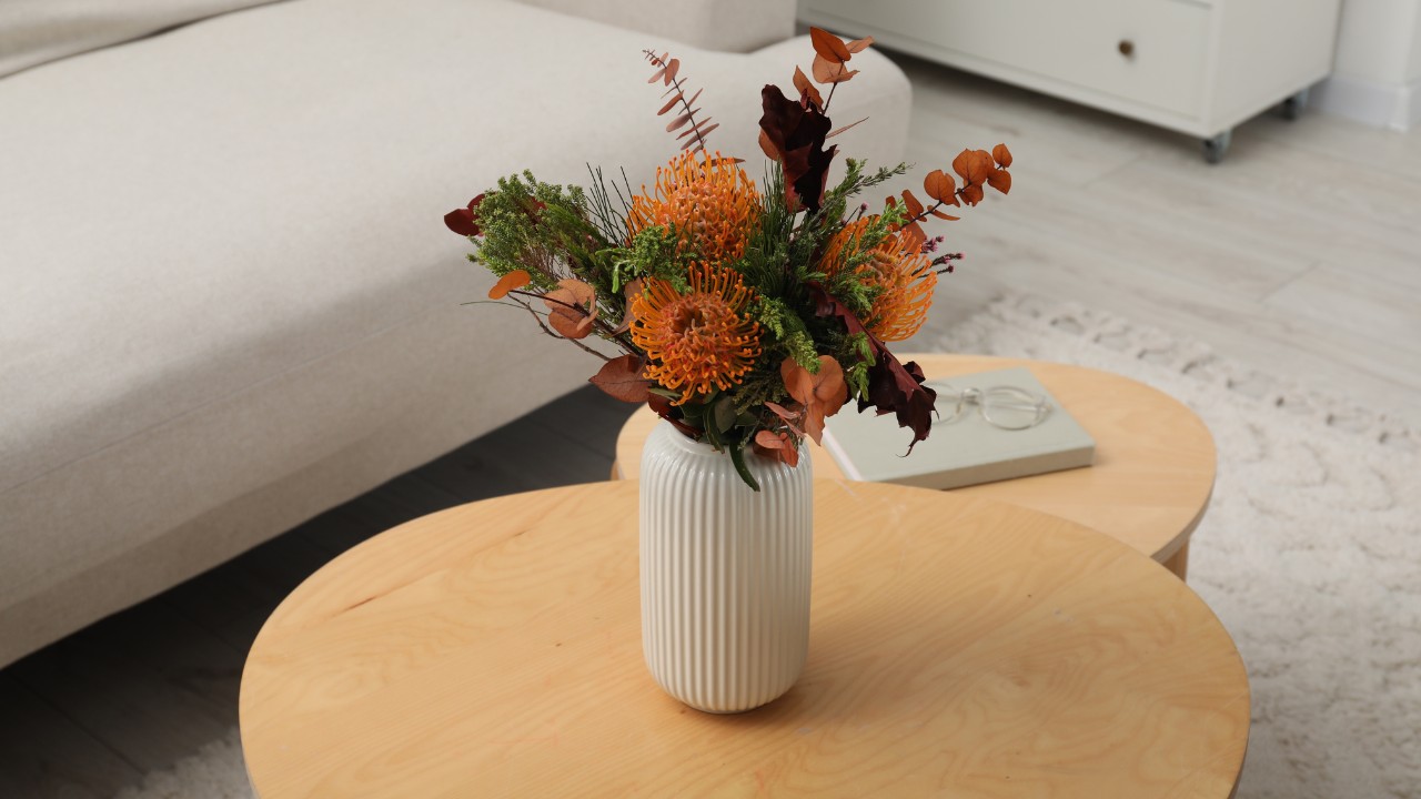 Vase with bouquet of beautiful leucospermum flowers, book and glasses on wooden nesting tables indoors.