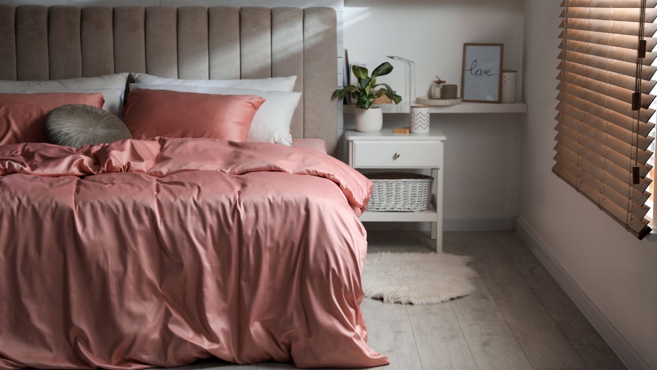 Bed with beautiful silky linens in stylish room interior. A white nightstand with plant and basket sit beside the bed in front of similarly-styled floating shelves. Natural light peers through the blinds in the window beside the shelves.