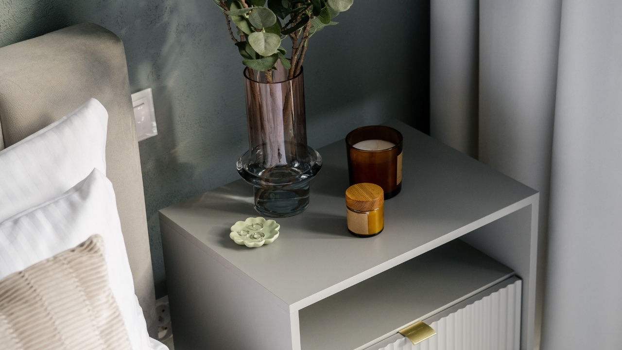 View of a nightstand in a bedroom, with plant in vase, candles, and jewelry dish.