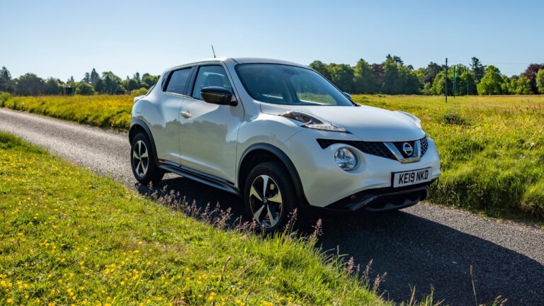Front-side view of a white Nissan Juke, driving on gravel by dandelion grass.