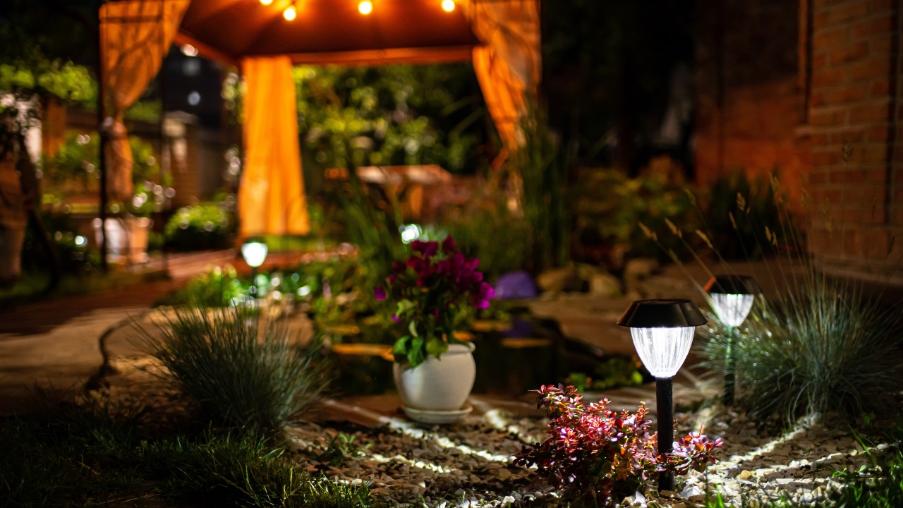 An outdoor assortment of greenery, plants, and decor surrounding a covered outdoor patio area. Stake lights and hanging Edison bulbs illuminate the area.