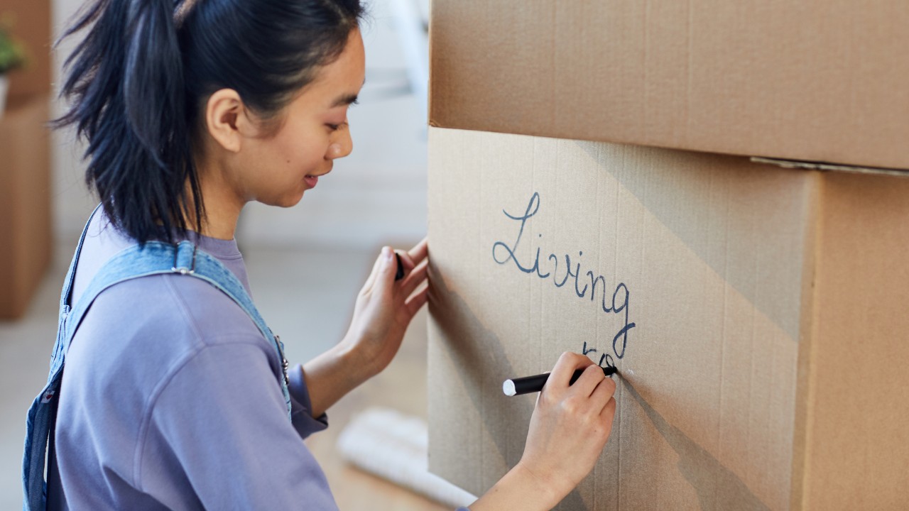 A young woman writing on stacked boxes with a permanent marker. The box is labeled "Living Room", and it appears they are decluttering, moving or selling goods. 