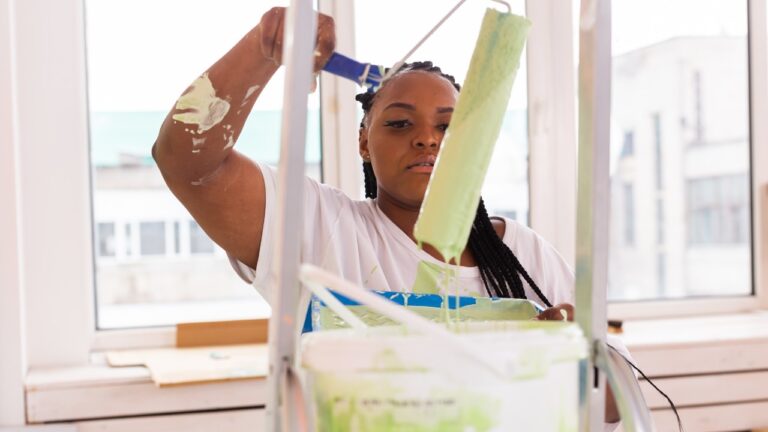 A woman lifting up a paint roller covered in light green paint, watching the runoff drip into the bucket.