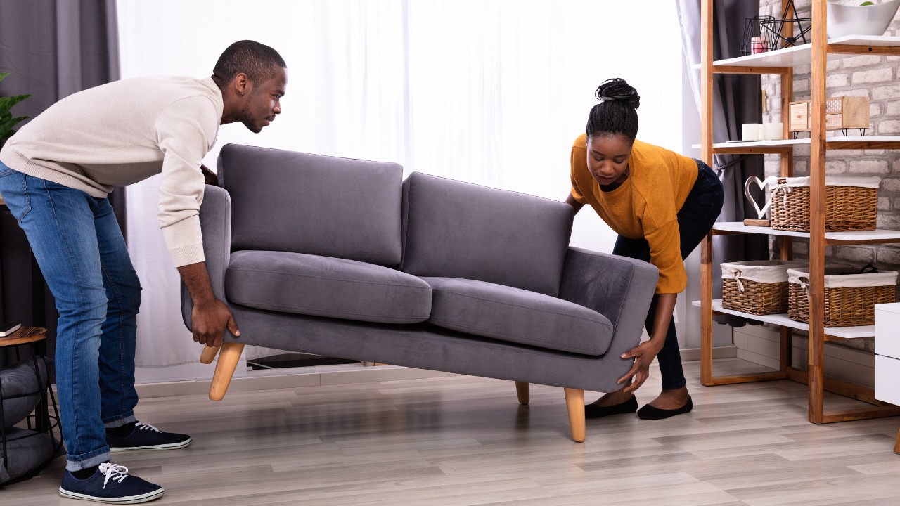A young couple moving a small grey couch together, one person at each end. 