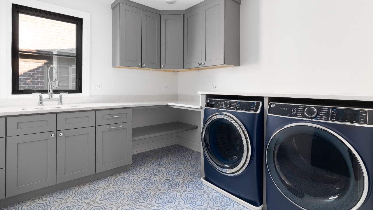 A beautiful laundry room with blue washer and dryer appliances, mosaic tiled floor, grey cabinets, and white marble countertop.