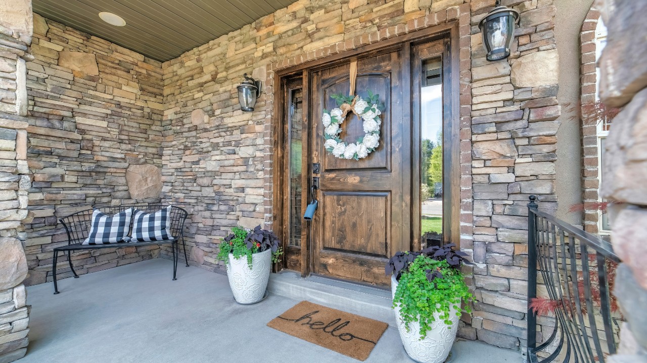 Entrance of a house with stone walls lockbox on the wooden front door with two sidelights, white wreath and two potted plants on the door and a bench on the left.