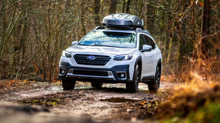 White Subaru Outback car with ski box drives on a muddy road in the forest.