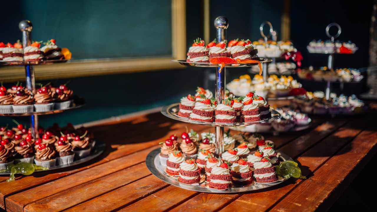 Tiered trays of assorted desserts, including cakes and cupcakes, displayed on a wooden table with a dark blue background.