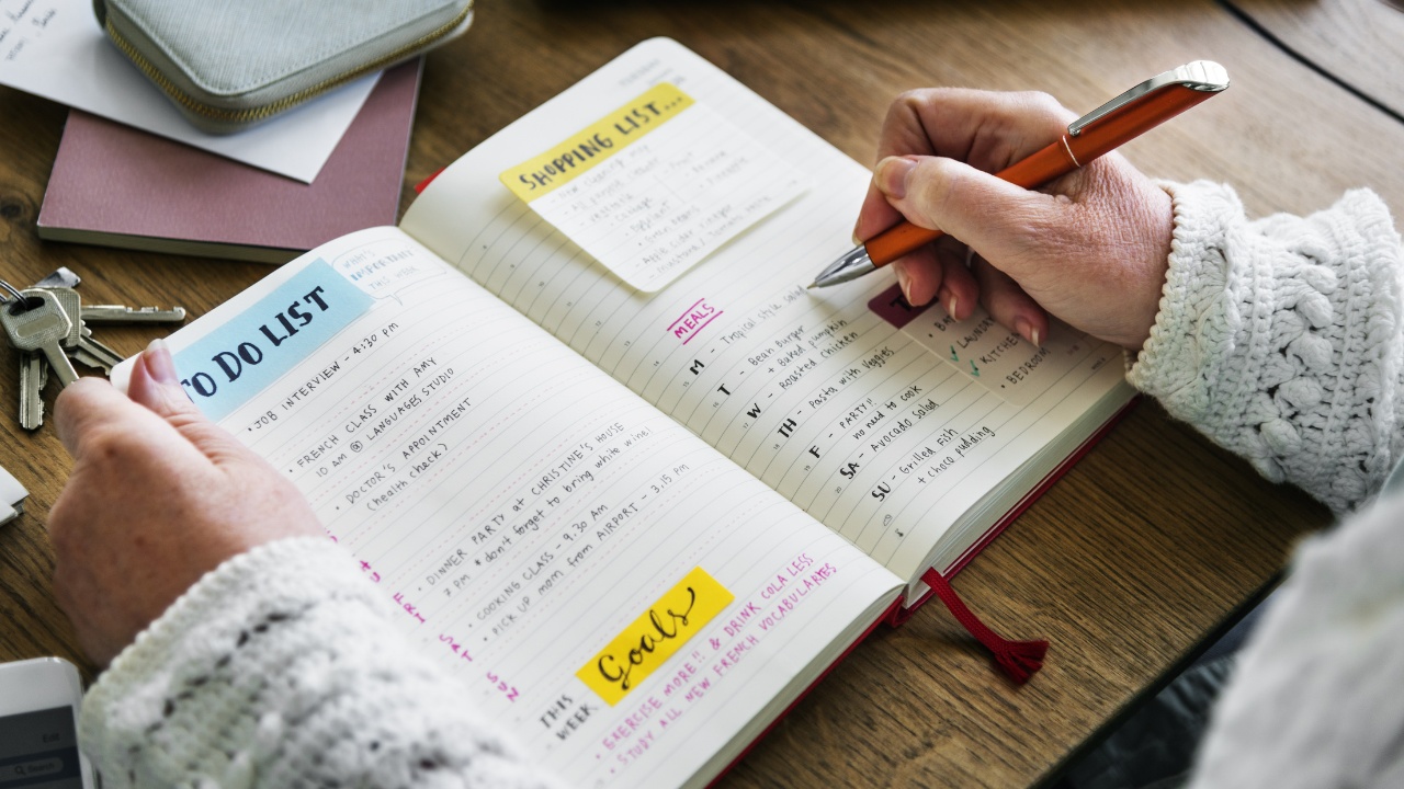 A senior woman writing to do lists, shopping lists and goals in a small journal. Her belongings are scattered around her on the tabletop, including a wallet and laptop.