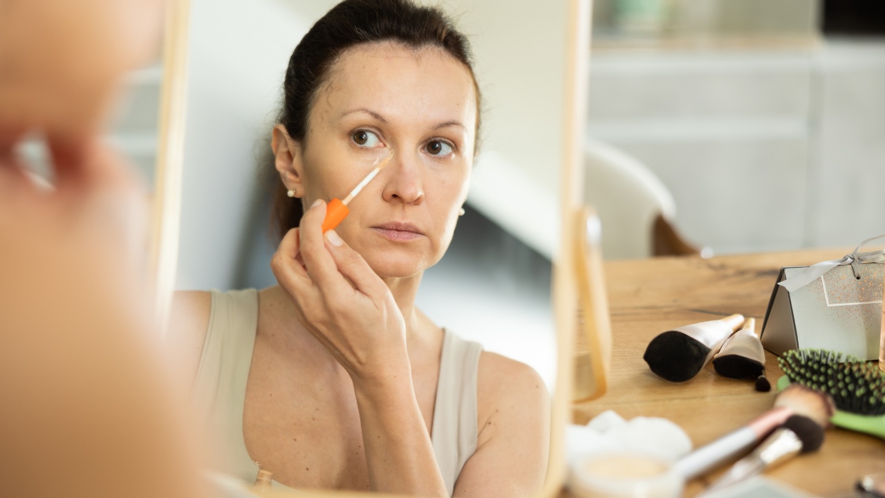 Careful middle-aged lady doing makeup with concealer while seated at a table in her house.