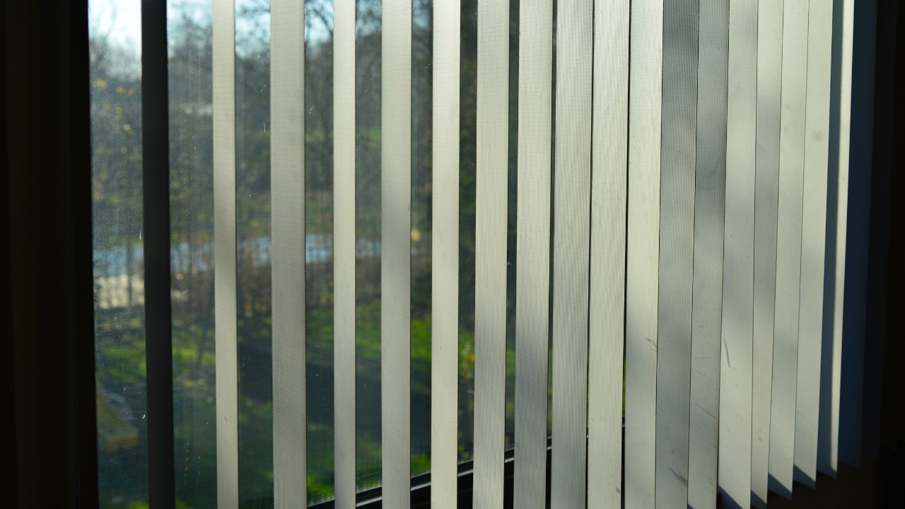 View from a bed angle to a sunlit window with a white plastic slatted curtain in a bedroom with shadows and shades of light on the slats.
